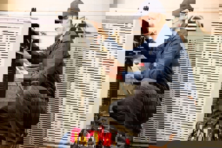 A technician kneels, inspecting an HVAC unit with a tablet.