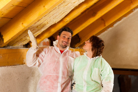 Here's an image of a couple in their attic, wearing coveralls, installing insulation between exposed wooden rafters.