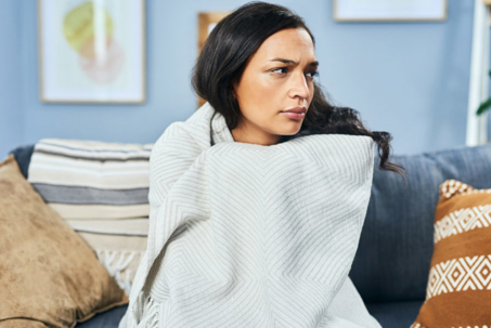A woman sits on a couch wrapped in a blanket looking upset