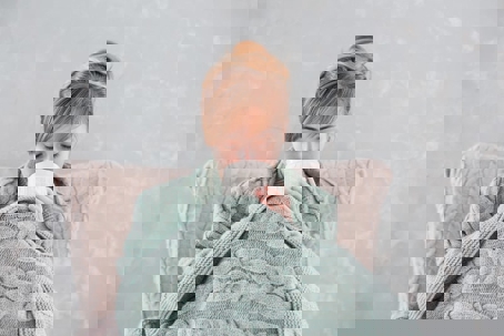 A woman wrapped in a cable-knit blanket sits and drinks from a mug.