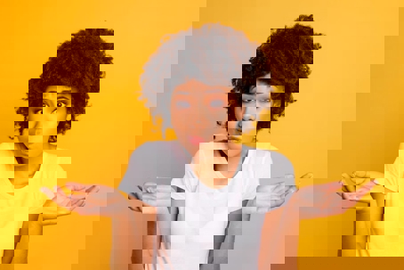 A woman with an afro and a white t-shirt stands against a yellow background