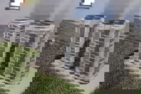 Two modern air conditioning units sit side by side on a concrete pad, with grass and the exterior of a building in the background.