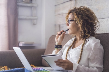 Woman with curly hair sits on a couch, holding a pen and notepad while looking at a laptop.