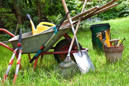 Various gardening tools such as a wheelbarrow, rakes, and shovel lay on a green lawn, ready for spring