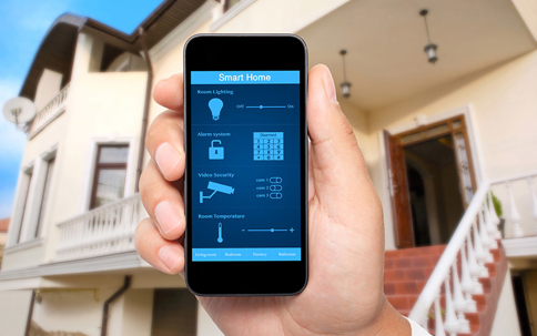 A hand holds a smartphone displaying a smart home app in front of a house.